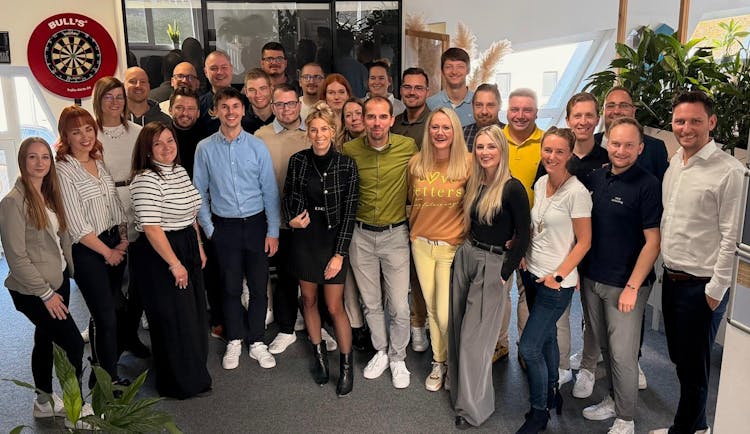 Gruppe von 27 Personen unterschiedlicher Geschlechter und Altersgruppen, die in einem Büro mit Pflanzen und Dartscheibe lachend zusammenstehen.